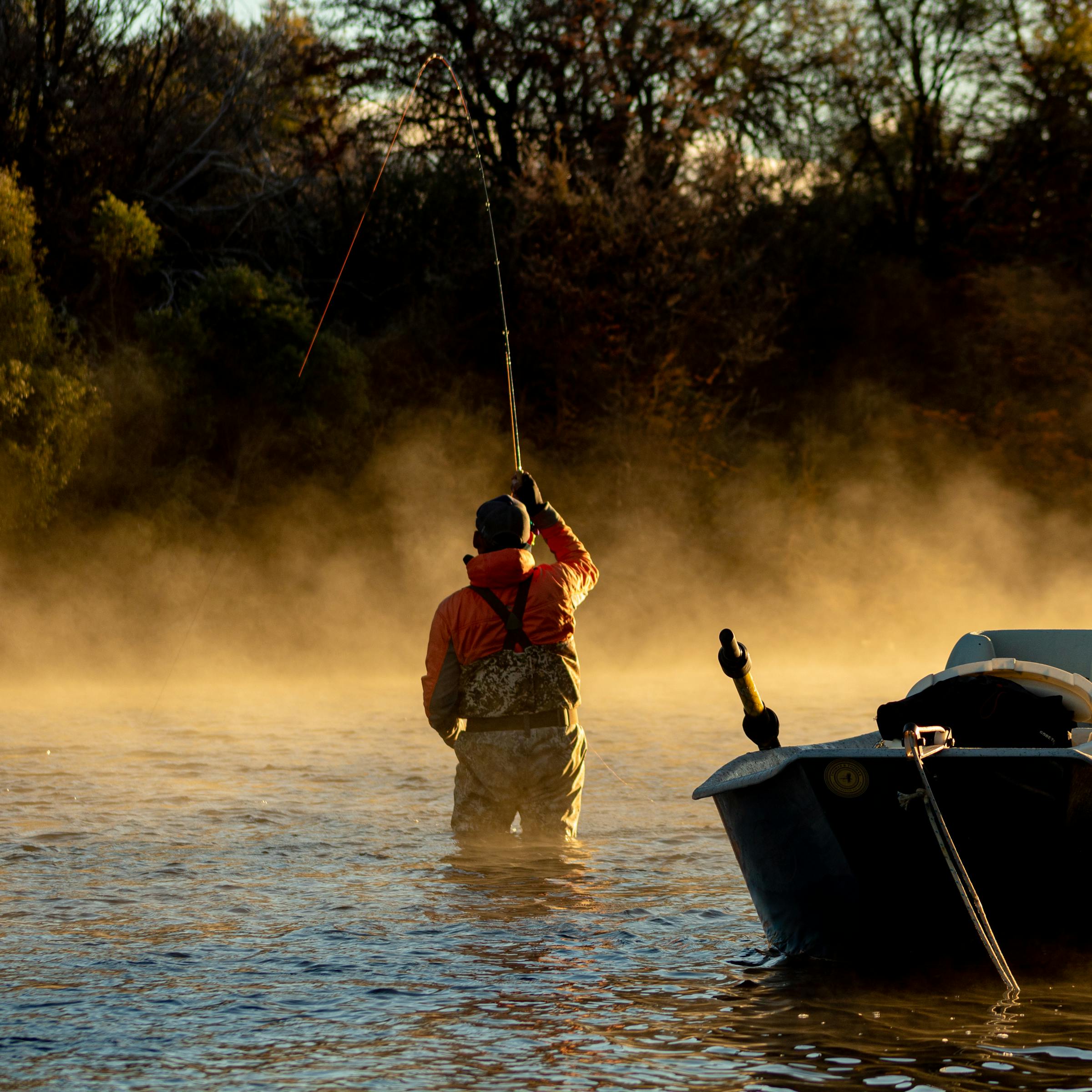 A fly fisherman in golden mist at dawn with a drift boat in the river